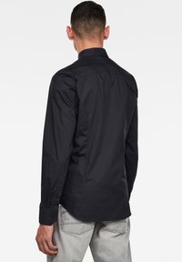 Camicia nera a maniche lunghe con colletto strutturato, realizzata in tessuto liscio, con polsini a bottone e una vestibilità sartoriale, mostrata dal retro.