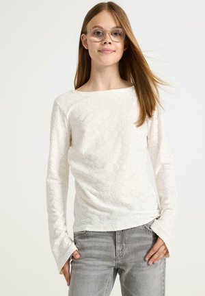 Haut blanc à manches longues avec un motif floral texturé, décolleté bateau large et manches évasées, associé à un jean gris clair.
