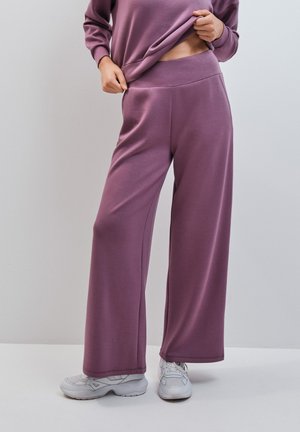 Personne portant un pantalon ample mauve et un haut assorti, relevant légèrement la chemise pour révéler la taille, avec des baskets blanches sur fond uni.
