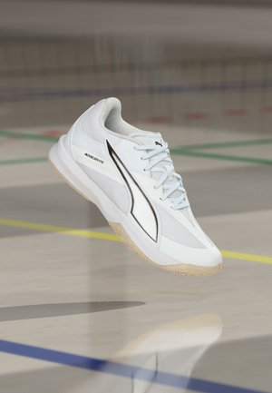 Bílá sportovní obuv Puma Accelerate s detaily z mesh, černým logem a světle gumovou podrážkou, zobrazena ve vzduchu na pozadí halového hřiště.