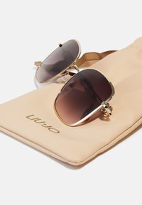 Gafas de sol con marco dorado y lentes oscuros con degradado, colocadas sobre un suave estuche beige que presenta el logo "Liu Jo" en dorado.