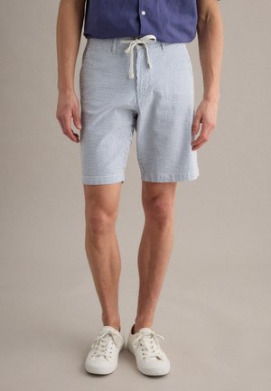 Mann trägt hellblau-weiß gestreifte Shorts mit Kordelzug, weiße Sneaker und ein dunkelblaues Hemd mit Knöpfen.