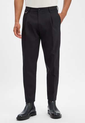 Pantalon noir ajusté slim-fit avec passants de ceinture, poches latérales et plis, porté avec des bottines en cuir noir.
