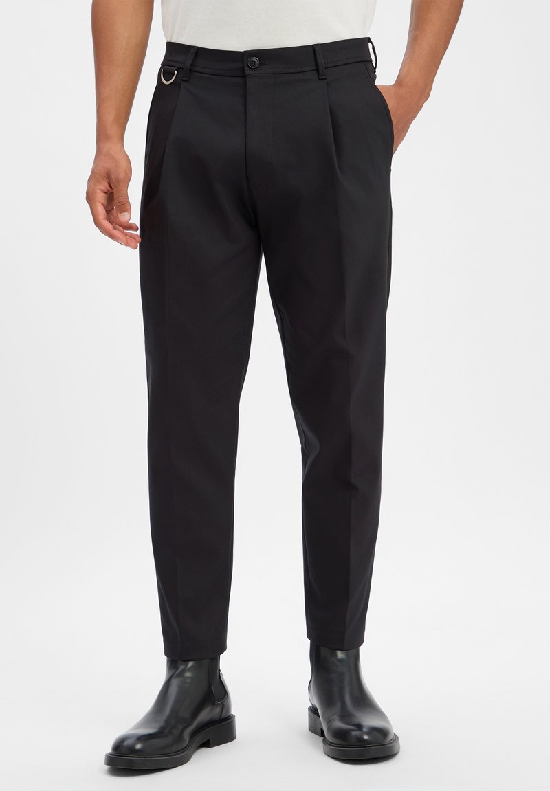 Pantalon noir ajusté slim-fit avec passants de ceinture, poches latérales et plis, porté avec des bottines en cuir noir.
