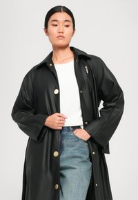 Cappotto impermeabile nero con bottoni dorati a scatto, tasca con zip e vestibilità rilassata. Indossato sopra una maglietta bianca e jeans blu. Texture liscia.