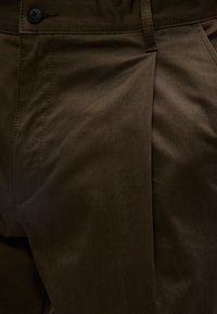 Κοντινό πλάνο σκούρου καφέ παντελονιού που δείχνει λεπτομέρειες όπως κουμπί, θηλιά ζώνης, τσακίσματα και πλαϊνή τσέπη.