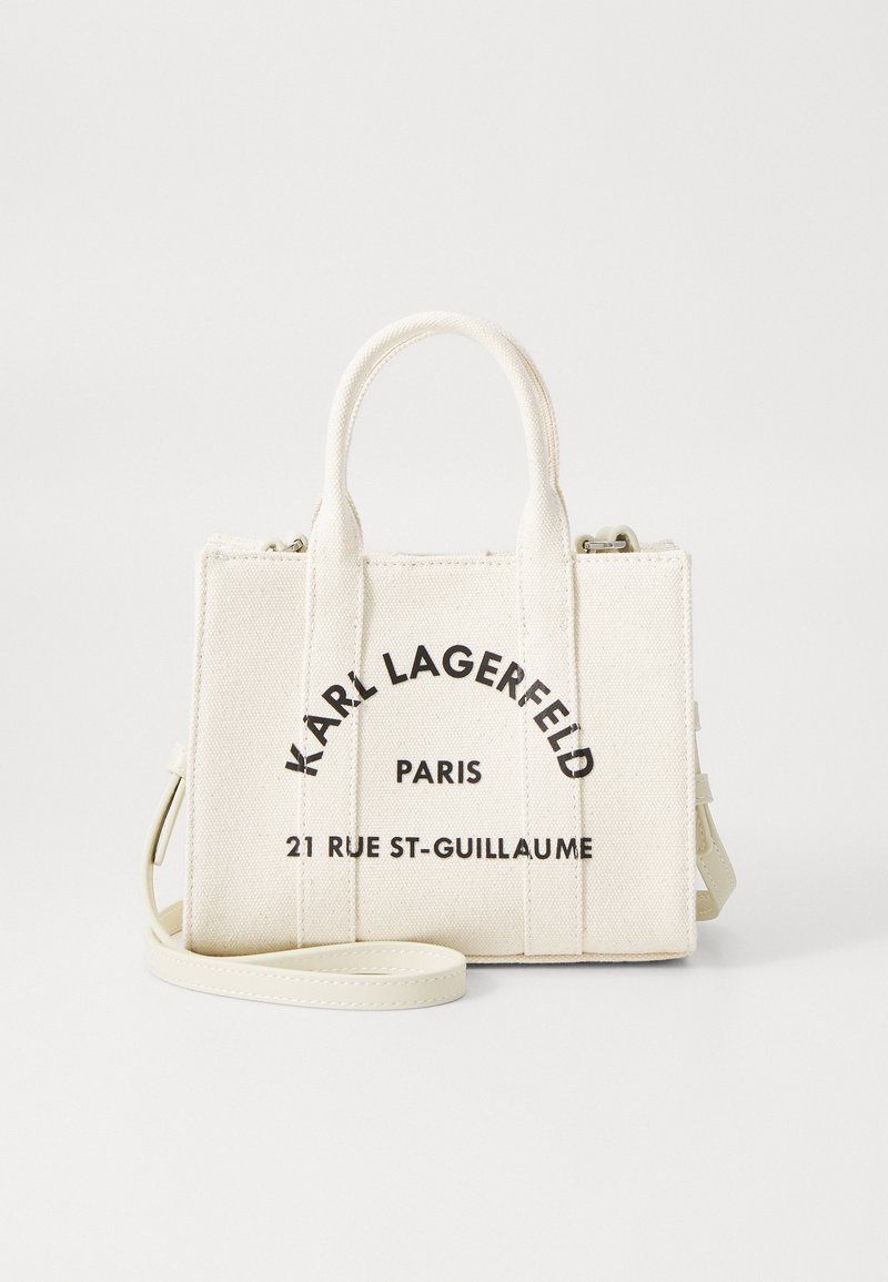 Balta audekla rokassoma ar melnu tekstu: "KARL LAGERFELD," "PARIS," un "21 RUE ST-GUILAUME." Ietver arī īsas rokturi un noņemamu siksnu.