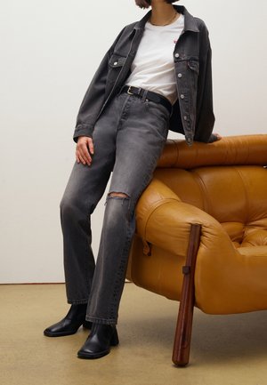 Veste en denim gris et jean, t-shirt blanc, ceinture noire et bottes à talons noires. Le mannequin s'appuie contre une chaise en cuir marron.