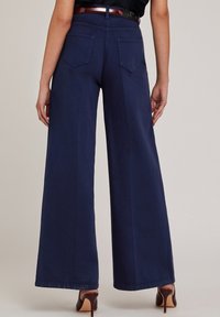 Marineblauwe wijde broek gemaakt van een gladde stof, met twee achterzakken en een hoge taille. De outfit wordt compleet gemaakt met bruine hakken.