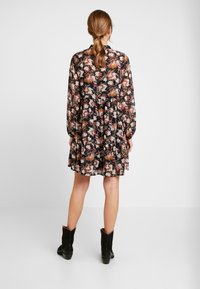 Femme avec des cheveux bruns en queue de cheval portant une robe à manches longues à fleurs et des bottes noires mi-mollet, se tenant devant un fond blanc uni.