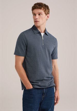 Polo azul marino con cuello de media cremallera, mangas cortas, tejido texturizado y bolsillo frontal, combinado con jeans azules. Diseño limpio.
