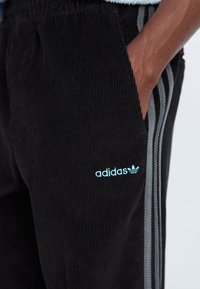 Black corduroy joggers featuring an elastic waistband, two side pockets, gray stripes along the sides, and a light blue Adidas logo.