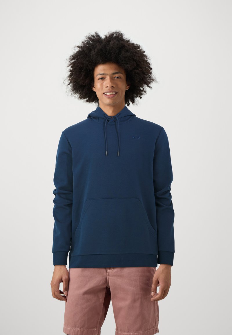 Sudadera azul marino con bolsillo canguro, capucha con cordón y puños acanalados, combinada con pantalones cortos rosa claro. Textura de tela suave.