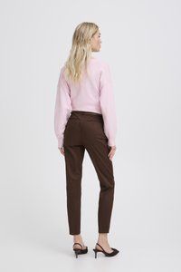 Sieviete ar gariem blondiem matiem, tērpusies rozā džemperī, brūnās piegulošās biksēs un melnās augstpapēžu sandales ar siksniņu, stāvot ar muguru pret skatītāju.