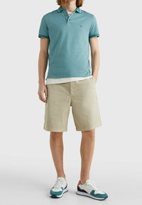 Muž v tyrkysovém polo tričku, béžových kraťasech ke kolenům a bílých teniskách s tyrkysovými a námořnickými akcenty, stojící s rukama v kapsách.