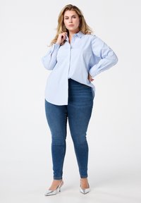 Chemise boutonnée à rayures bleu clair et blanche, coupe décontractée, manches longues, assortie à un jean skinny bleu foncé et des escarpins argentés.
