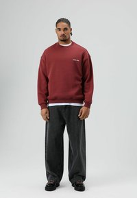 Sweatshirt bordeaux à col rond, logo blanc sur la poitrine, associé à un jean noir ample et des chaussures noires. Texture lisse, design décontracté.