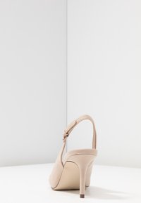 Escarpin beige à talons hauts avec une texture en daim, dos ouvert et bride de cheville. Le talon est fin et mesure environ 7,5 cm de hauteur.