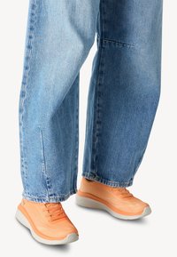 Orange running shoes with a textured upper and grey sole, paired with loose fitting light blue denim jeans.