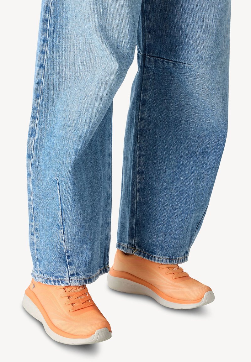 Orange running shoes with a textured upper and grey sole, paired with loose fitting light blue denim jeans.