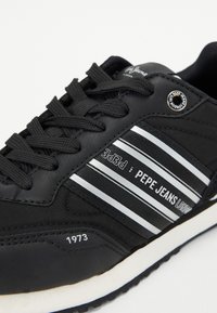 Scarpa sportiva nera realizzata in materiale sintetico, con strisce bianche, dettaglio del logo e lacci testurizzati. La suola è bianca con una presa testurizzata.