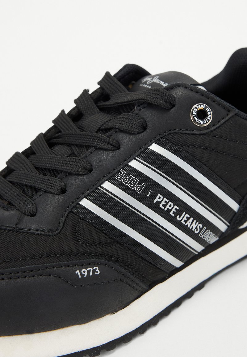 Chaussure de sport noire en matériau synthétique, avec des rayures blanches, un logo et des lacets texturés. La semelle est blanche avec une prise texturée.
