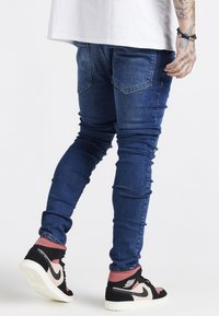 Schmale blaue Jeans mit abgenutzten Akzenten, zwei Gesäßtaschen, kombiniert mit schwarzen und pinken High-Top-Sneakers.