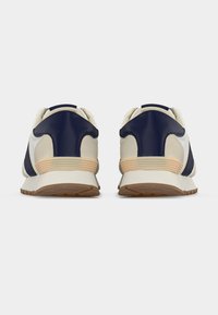 Ζευγάρι λευκά και ναυτικά αθλητικά παπούτσια με μπεζ λεπτομέρειες, εμφανιζόμενα από πίσω σε λευκό φόντο.