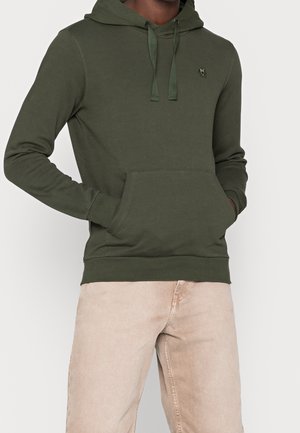 Homme portant un sweat à capuche vert olive avec une poche avant et un pantalon beige, les mains glissées dans la poche.