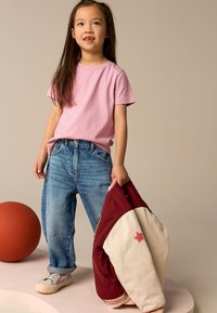 Maglietta di cotone rosa chiaro, jeans blu, tiene una giacca bomber bordeaux e crema con una toppa a forma di stella, e sneakers multicolore. Pallone arancione sullo sfondo.