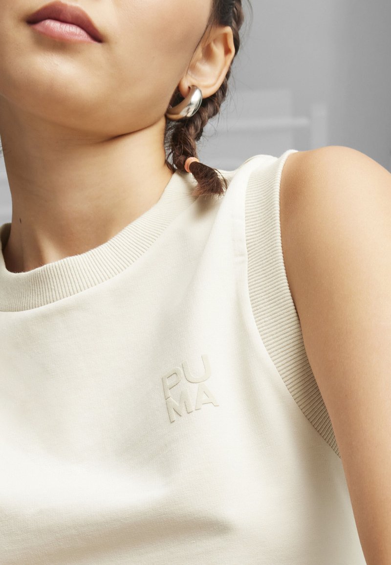 Jasno-beżowy bezrękawnik z miękkiego materiału, z karczkiem w prążek. Posiada wytłoczone logo "PUMA" na froncie, subtelny i minimalistyczny design.