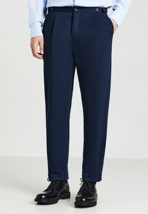 Homme portant un pantalon bleu marine sur mesure, des chaussures habillées en cuir noir et une chemise button-up bleu clair avec une main dans sa poche.