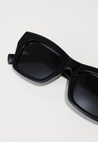 Ochelari de soare negri, supradimensionați, cu un cadru gros, unghiular și lentile întunecate. Suprafață netedă cu o finisare mată. Fără branding sau modele vizibile.