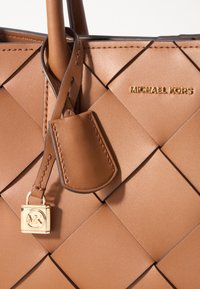 Bolso de piel marrón claro con diseño trenzado, presenta el logo de Michael Kors en dorado, una etiqueta colgante y un cierre dorado. Textura suave y forma estructurada.
