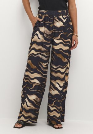 Vrouw in losse, high-waisted broek met zwarte, beige en bruine golvende abstracte patronen, gecombineerd met zwarte open teen sandalen.