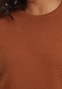 Pletený svetr v teplé hnědé barvě s texturovaným vzorem včelí plástve a žebrovaným kulatým výstřihem. Detailní pohled na tkaninu.
