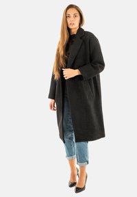 Donna con capelli lunghi che indossa un lungo cappotto nero, jeans blu arrotolati e scarpe con tacco alto nere, in piedi su uno sfondo chiaro.