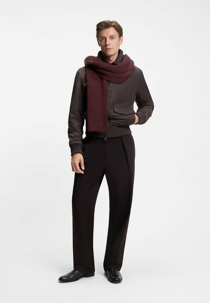 Brun bomberjakke med ribbede manchetter, parret med et maroon ribbet tørklæde og sorte bukser. Modellen står foran en neutral baggrund.