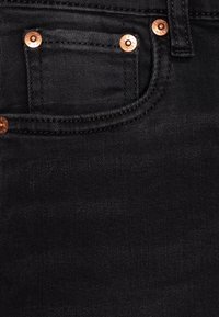 Svarta denimjeans med en närbild av fickområdet, som har kopparfärgad detaljer, kontrasterande sömmar och en slät yta.