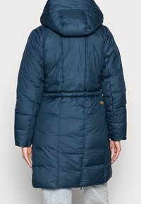 Mörkblå puffjacka med huva, åtdragen midja, långa ärmar och quiltad textur; har en liten logotyp på nedre delen av ryggen.