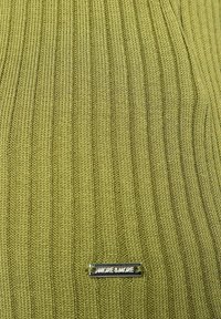 Nærbillede af ribbet olivengrøn strikstof med et lille rektangulært metallisk mærke med teksten "MORE & MORE", syet nær bunden i midten.