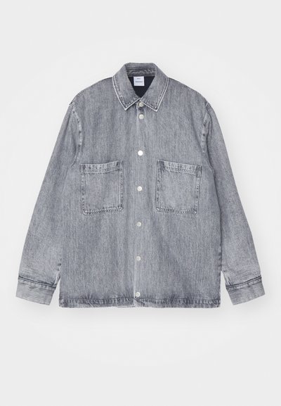 Veste en denim gris clair, avec un devant boutonné, un col et deux grandes poches avant. Tissu texturé avec des détails de couture visibles.