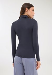 Kobieta w dopasowanej, granatowej koszulce z długim rękawem i luźnych, jasnoszarych spodniach, stojąca przodem z odwróconą głową w prawo.