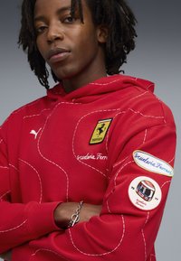 Rode hoodie met een gestikt ontwerp, Scuderia Ferrari-embleem en een patch met een racehelm graphic. De stof heeft een gladde textuur.