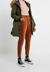 Zelená parka s kožešinovou kapucí, růžový žebrovaný rolák, hnědé kordové kalhoty a černé vysoké tenisky. Neformální outfit vhodný pro chladné počasí.
