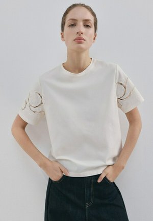 Mladá žena v bielom krátkornom tričku s kvetinovou čipkovanou výšivkou na rukávoch a tmavých vysokých nohaviciach, stojaca s rukami v vreckách.