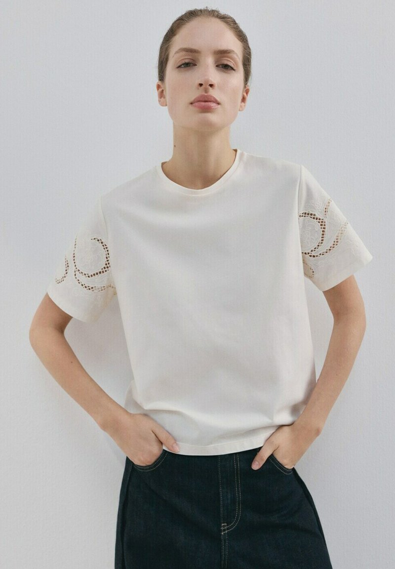 Mladá žena v bielom krátkornom tričku s kvetinovou čipkovanou výšivkou na rukávoch a tmavých vysokých nohaviciach, stojaca s rukami v vreckách.