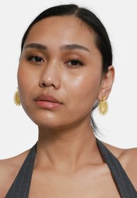Pendientes en tono dorado con un diseño de estallido solar, que presentan rayos texturizados y un centro circular. La modelo lleva un top de halter oscuro con rayas.