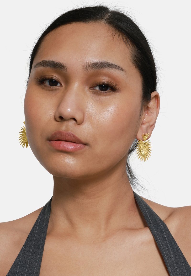 Pendientes en tono dorado con un diseño de estallido solar, que presentan rayos texturizados y un centro circular. La modelo lleva un top de halter oscuro con rayas.