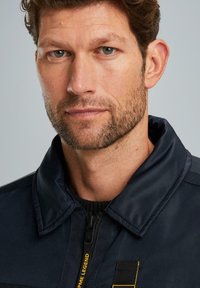 Marineblaue Jacke mit einem Reißverschluss vorne, hohem Kragen und strukturiertem Stoff. Mit gelbem Logoakzent und robusten Nähdetails.